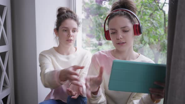 Slim Attractive Caucasian Young Woman in Headphones Enjoying Music As Twin Sister Asking Tablet