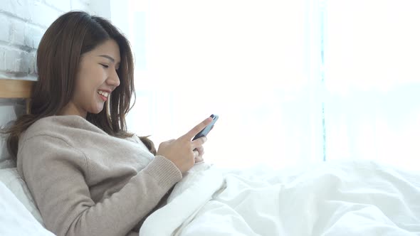 Asian woman in bed checking social apps with smartphone.