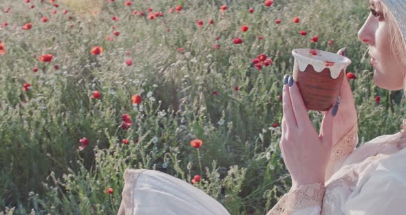 A Young Woman in a White Dress and a Hat Drinks Tea Against the Backdrop of a Wild Field in the Rays