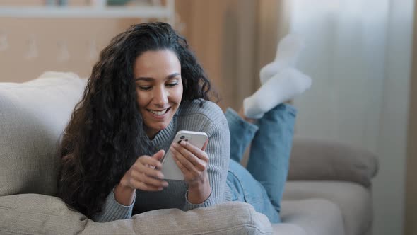 Happy Hispanic Young Woman Lying on Sofa Typing Message on Phone Smiling Carefree Girl Communicate
