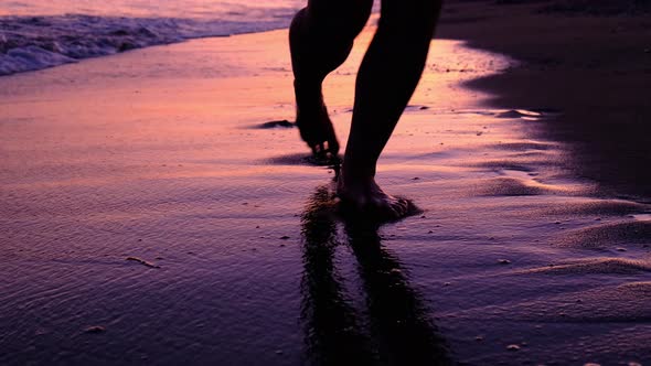 Female Legs and Feet Walking Along Sea Water Waves on Sandy Beach on Pink Sunset