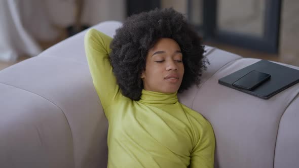 Young Smiling African American Woman Lying on Couch Dreaming with Smartphone and Tablet Aside