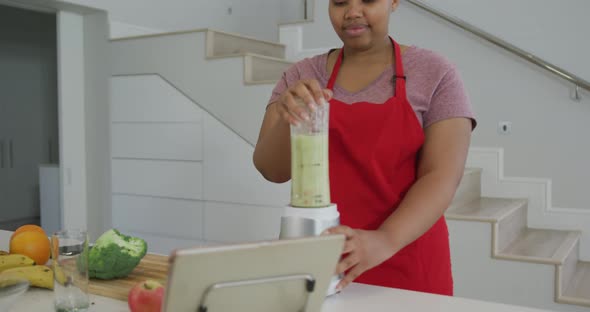 Happy african american plus size woman wearing apron, preparing health drink in kitchen