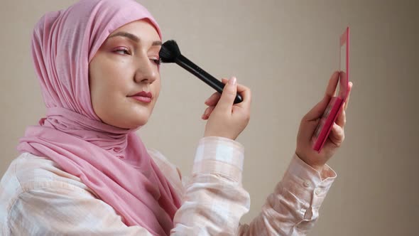 Young Muslim Woman in Hijab Applying Makeup with a Brush Looking in the Mirror