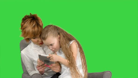 Young Mother and Her Daughter Sit Together and Look at the Tablet on a Green Screen, Chroma Key