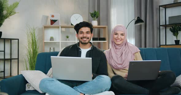 Multiethnic Couple Sitting in Relaxed Pose with Computers on Comfortable Couch at Home