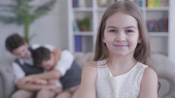 Joyful Little Brunette Girl Smiling at Camera and Running To Twin Brothers Playing on Couch 