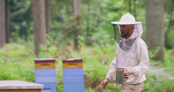 Bearded Young Male Beekeeper in White Protective Suit Walks Through the Apiary Between the Hives in