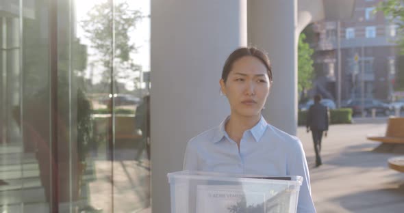 Slider Shot of Asian Sad Businesswoman Walking Outdoors with Box of Stuff Leaving Business Center