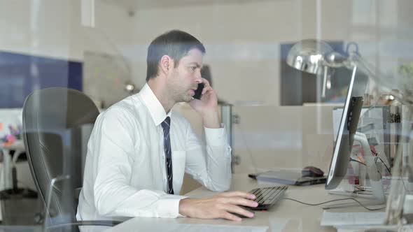 Angry Middle Aged Businessman Talking on His Cellphone and Using Office Computer