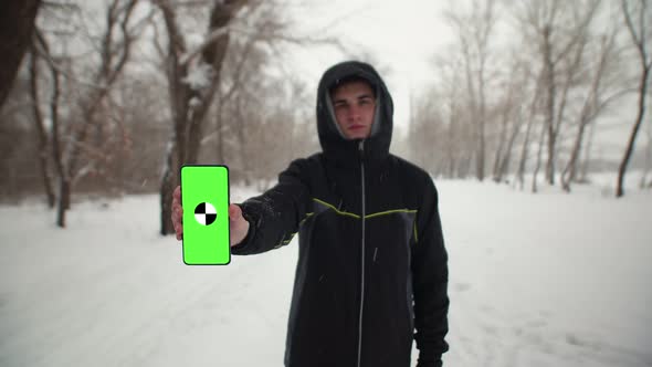 Caucasian Man in Black Jacket with Hood is Standing on Winter Street and Holding Phone with Green