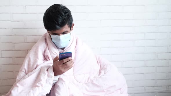 Young Man in Surgical Face Mask Using Smart Phone on Bed