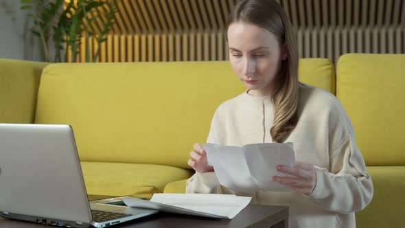 Young Woman Sits on the Floor Next To a Yellow Sofa at a Table with Paper Bills Feeling Stressed