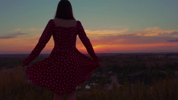 Rear view of woman in red dress swinging, standing on hill at sunset.