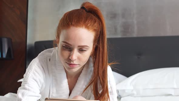 Young Redhead Woman Watching Movie on Tablet Computer Lying on Bed in Hotel Room