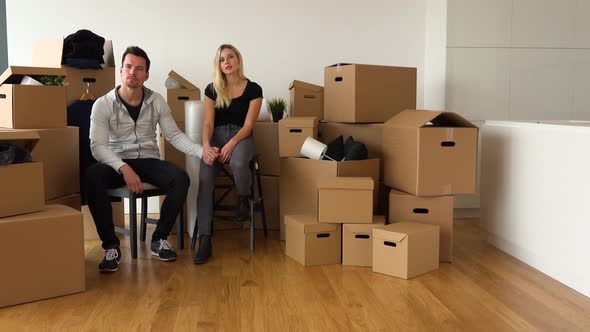 A Moving Couple Sits on Chairs, Holds Hands and Looks Seriously at the Camera in an Empty Apartment