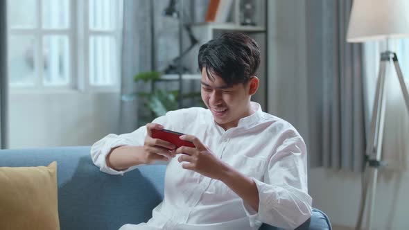 Asian Man Playing Game On Smartphone And Smiling While Lying On Sofa In The Living Room