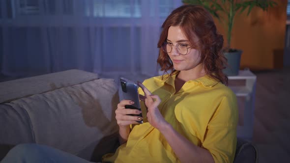 Woman with Glasses Uses Mobile Phone and Internet While Relaxing on Sofa in Living Room in Evening