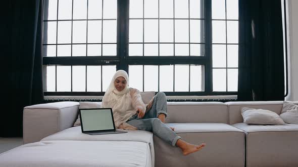 Young Muslim Girl Sits on Couch and Looks at the Laptop in Bright Beige Studio