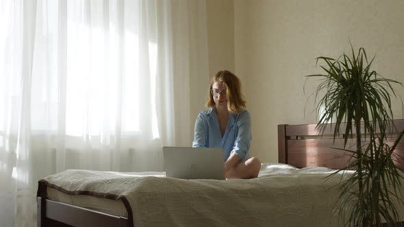 Woman freelancer sitting on bed uses laptop. A woman works or studies remotely.