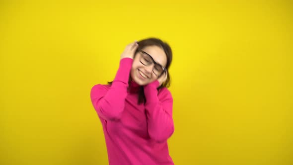 Young Woman Dancing with Headphones on a Yellow Background. A Girl in a Pink Turtleneck and Glasses