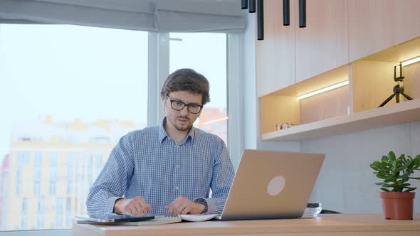 Young man is stretching at the computer, raising his hands up, resting from the work done.