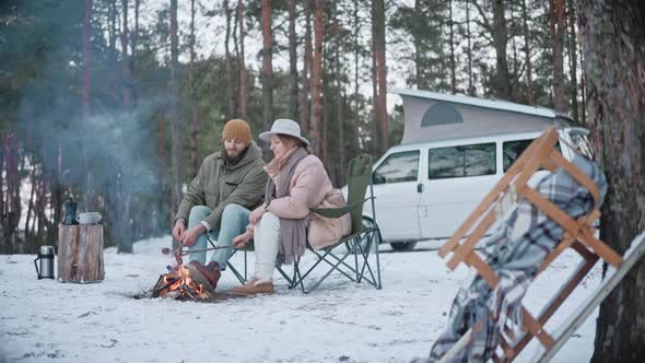 A Couple is Roasting Sausages on Sticks on a Campfire in the Woods in Winter