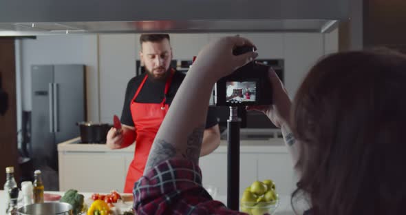 Woman with Camera Filming Man Chopping Vegetables in Kitchen for Food Blog