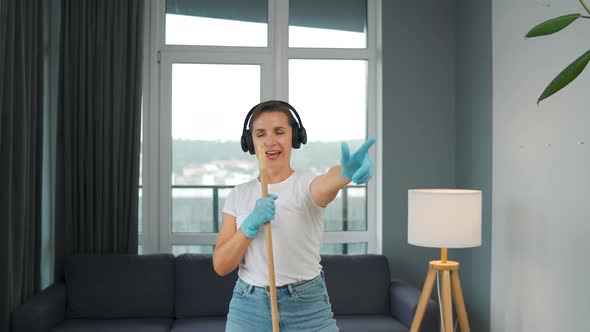 Woman in Headphones Cleans the House and Have Fun Singing with a Broom Like a Star at a Concert