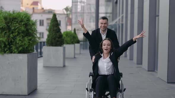 Cheerful Man Pushing Wheelchair with His Disabled Coworker and Smiling Near Office Building