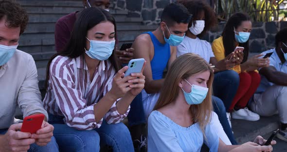 Multiracial young people having fun using mobile phone in the city while wearing safety face mask