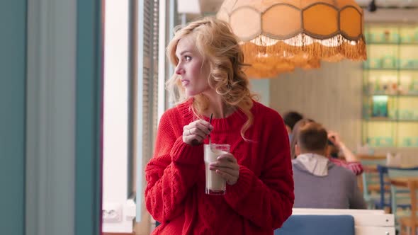 Beautiful Young Woman in a Red Sweater Drinks Coffee From a Straw in a Cafe. Blonde in a Stylish