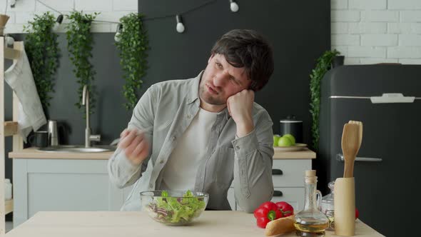 Unhappy Man Eating Vegetable Salad at Table in Kitchen