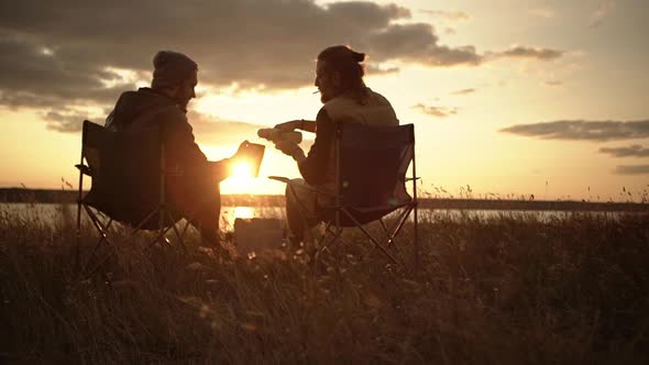 Two Men Sitting in Camping Chairs in a Field Watching Sunset Pouring Tea From Thermal Mugs
