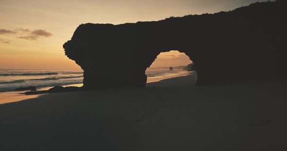 Slow Motion Sun Silhouette of Rock Wall with Giant Hole on Sand Beach Aerial View