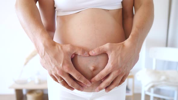 Hands Of Pregnant Woman And Her Husband Embracing Her Belly In Shape Of Heart.