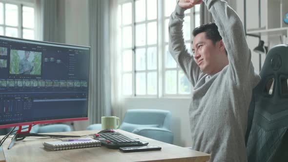 Asian Video Editor Man Stretching And Leaning Back While Using Desktop Computer At Home