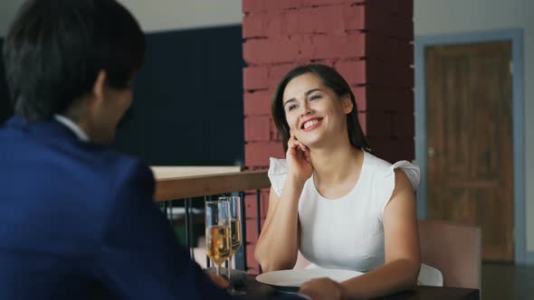 Happy Young Woman in Beautiful Dress Is Talking To Her Boyfriend on Date Sitting at Table Together