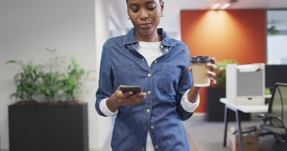 Video of african american businesswoman walking, drinking coffee and using smartphone in office