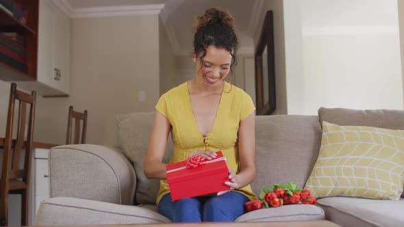 Happy biracial woman sitting on sofa, having valentines video call and opening present