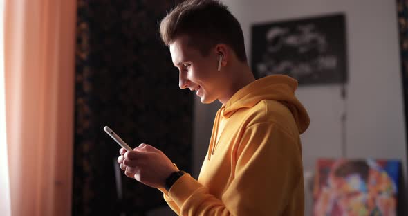 Man in Yellow Hoodie Using Smartphone and Earbuds at Home