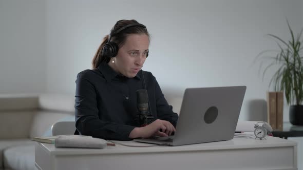 Young woman in a blue shirt sits in headphones and plays online game on laptop at home