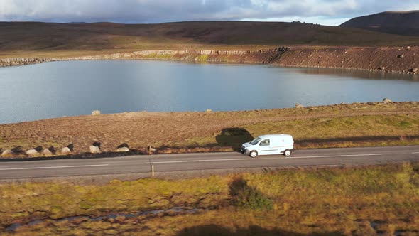 Profile shot of white car traveling on rural road along lake with bright sunlight, dusk