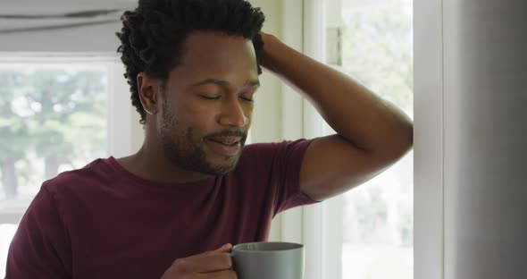 Portrait of relaxed biracial man standing at window with coffee and looking into distance