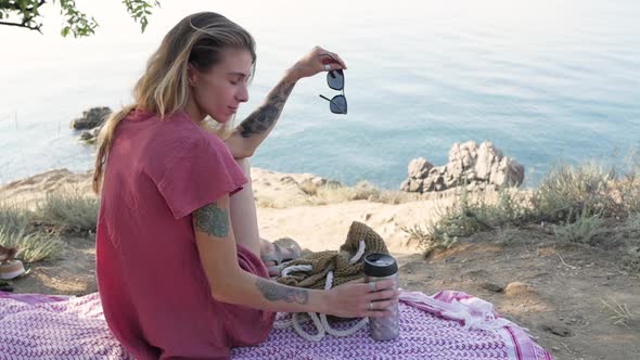 Young Woman Sitting on a Blanket on the Beach By the Sea Looking in Front of Her and Holding a