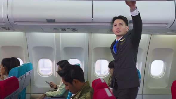 Caucasian male flight attendant walking on aislel to close luggage compartment in airplane.