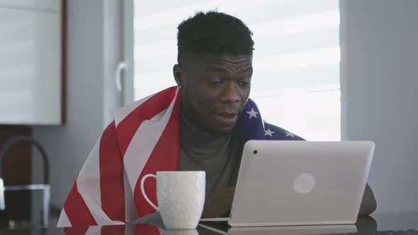 African American Man with USA Flag Over His Shoulders Watching Game on His Laptop with Cup of Tea on