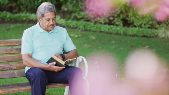 Video of relaxed biracial senior man reading book and sitting on bench in garden