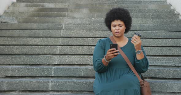 Happy plus size biracial woman using smartphone and drinking coffee