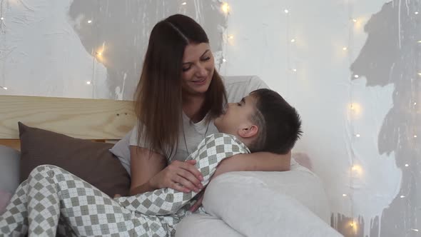 A Beautiful Mother Plays with Her Son Sitting on the Bed and Kisses Him Having a Good Mood